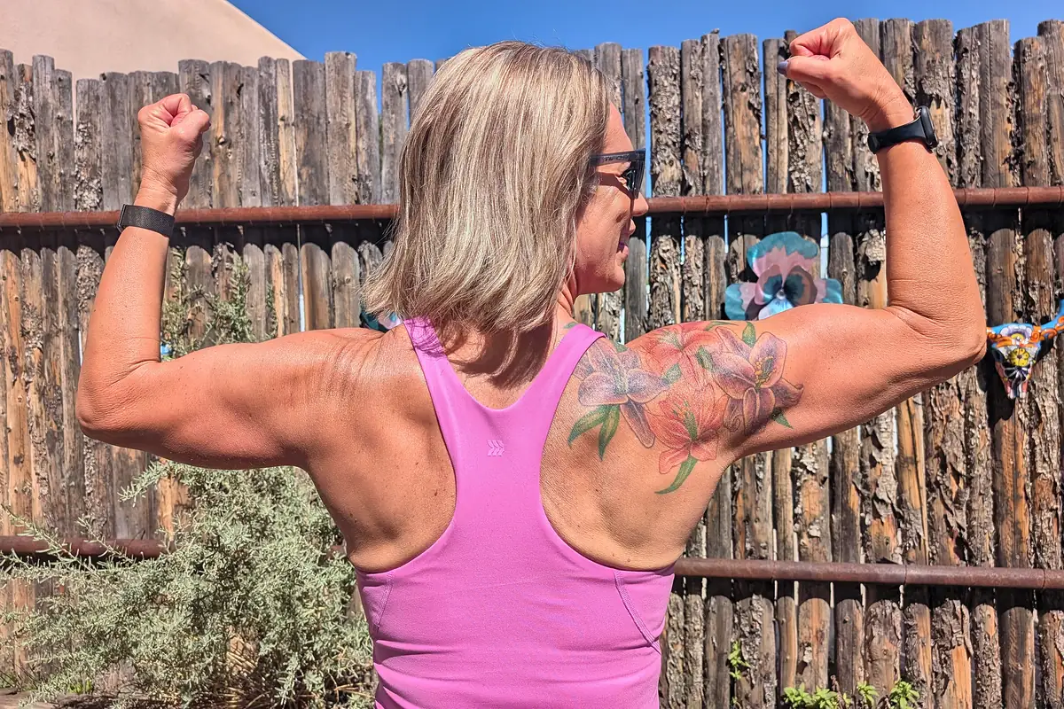 Jill flexing her arms, seen from behind, showing her shoulders, back and a floral tattoo