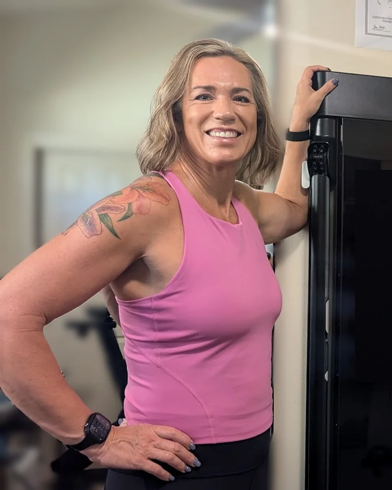 Jill McCracken smiling with one hand on her hip, resting her arm on a strength-training machine in her home gym