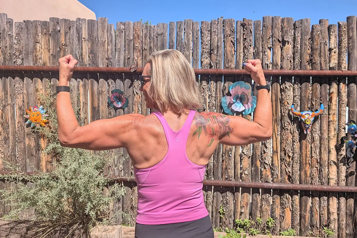 Jill from behind flexing both arms, showing her back and shoulders, in front of a wooden fence