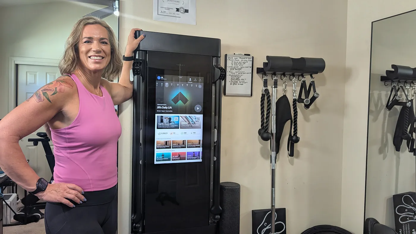Jill smiling in her home gym next to her strength-training machine, with her ISSA personal trainer certificate on the wall
