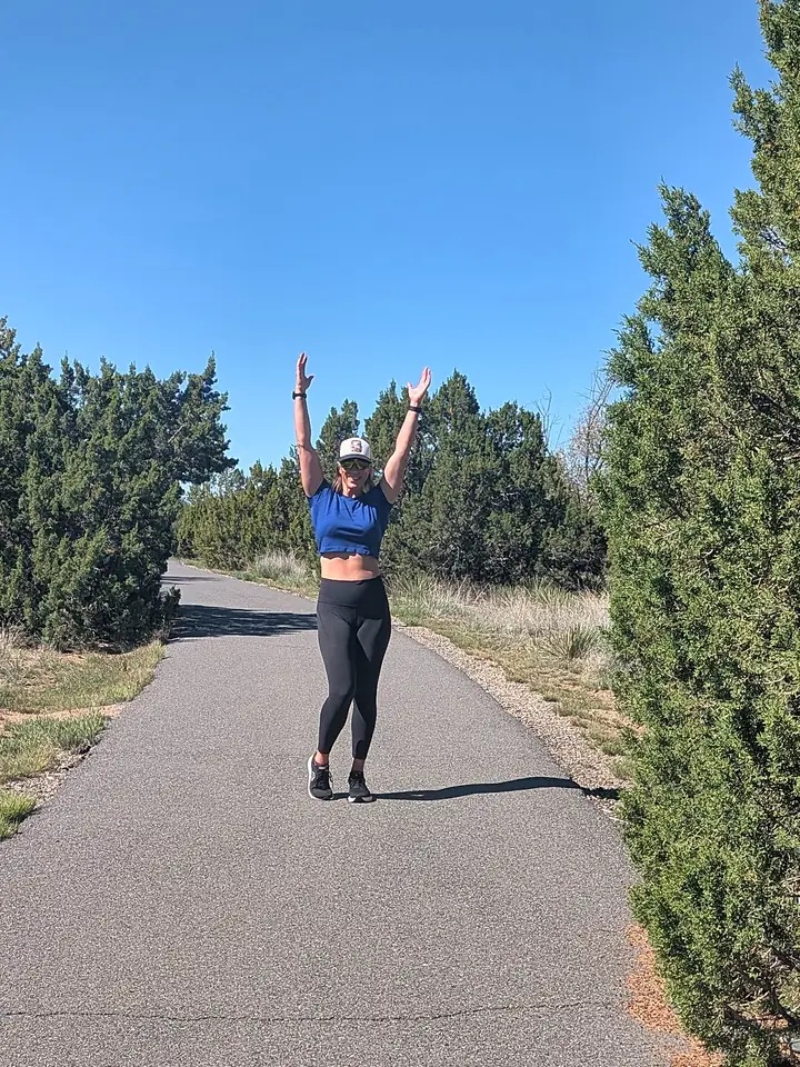 Jill on a paved trail between juniper trees under a blue sky, both arms raised